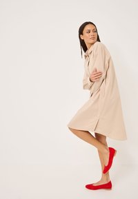 Abito camicia beige oversize con maniche lunghe, chiusura a bottoni e spacchi laterali, abbinato a ballerine in suede rosse.