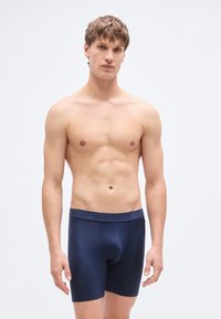 Μποξεράκια σε ναυτικό μπλε χρώμα, κατασκευασμένα από λείο ύφασμα με ελαστική μέση που φέρει ένα λογότυπο. Εφαρμοστό σχέδιο με μήκος μέχρι το μέσο του μηρού.