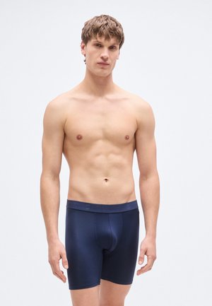 Tamsiai mėlyni bokseriai iš lygaus audinio su elastine juosmens juosta, puošta logotipu. Liesas dizainas, uždengiantis vidurį šlaunies.