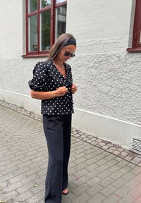 Kvinna i svart blus med prickar, svarta vida byxor, svart hårband och solglasögon, som justerar sin blus nära en texturerad vit vägg.