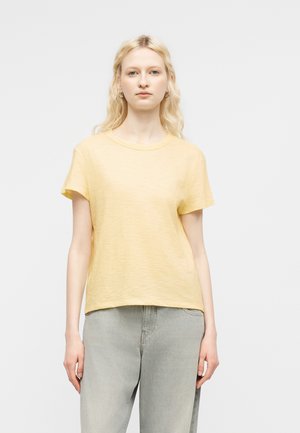 Młoda kobieta z długimi blond włosami, ubrana w jasnżółtą koszulkę z krótkim rękawem i jasnoszare jeansy, stojąca na tle jednolitej białej ściany.