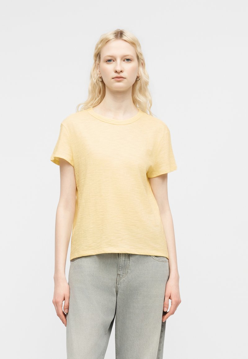 Νεαρή γυναίκα με μακριά ξανθά μαλλιά που φοράει ανοιχτό κίτρινο κοντομάνικο μπλουζάκι και ανοιχτό γκρι τζιν, στέκεται μπροστά σε απλό λευκό φόντο.