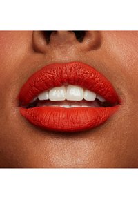 Klart röd matt läppstift på fylliga läppar, som visar upp en fin textur och slät yta mot en varm hudton med vita tänder synliga.
