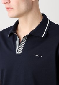 Polo shirt azul marino con cuello a rayas, con un diseño de cuello en V. Logo "GANT" en blanco en la parte delantera. Confeccionado en tela texturizada.