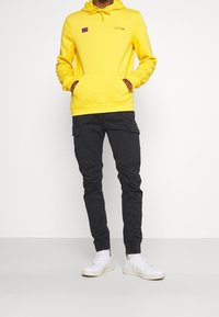 Geel hoodie met een voorzak en trekkoord, gecombineerd met zwarte cargobroeken met zijzakken en witte sneakers.