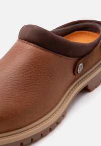 Brun lær slip-on træsko med polstret mørk brun krage, teksturert gummisåle og metall-logo på siden.