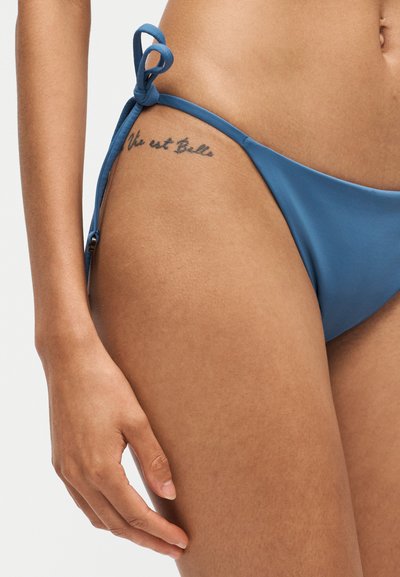 Gros plan sur la hanche et la main d'une personne portant un bas de bikini bleu avec un côté noué et un petit tatouage sur la hanche affichant "Vie est Belle."