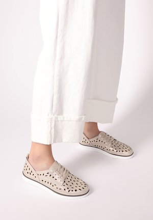 Beige perforated shoes with a textured surface, laces, and a white rubber sole, paired with loose white pants.