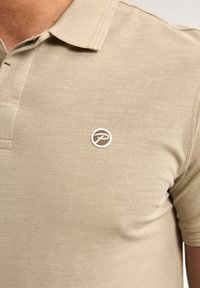 Béžové polo tričko s texturovanou látkou a třemi knoflíky u límce. Obsahuje kruhové hnědé logo na levé části hrudi.