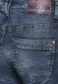 Jean en denim bleu foncé avec une texture lisse, présentant des coutures visibles, une poche arrière et une étiquette avec l'inscription "DENIM NE ME QUITTE JAMAIS CECIL."
