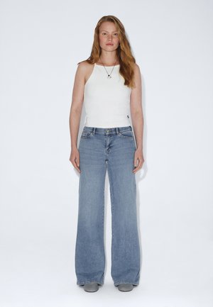 Vrouw met lang rood haar die een witte mouwloze top, wijde blauwe spijkerbroek en grijze schoenen draagt, staand tegen een effen witte achtergrond.