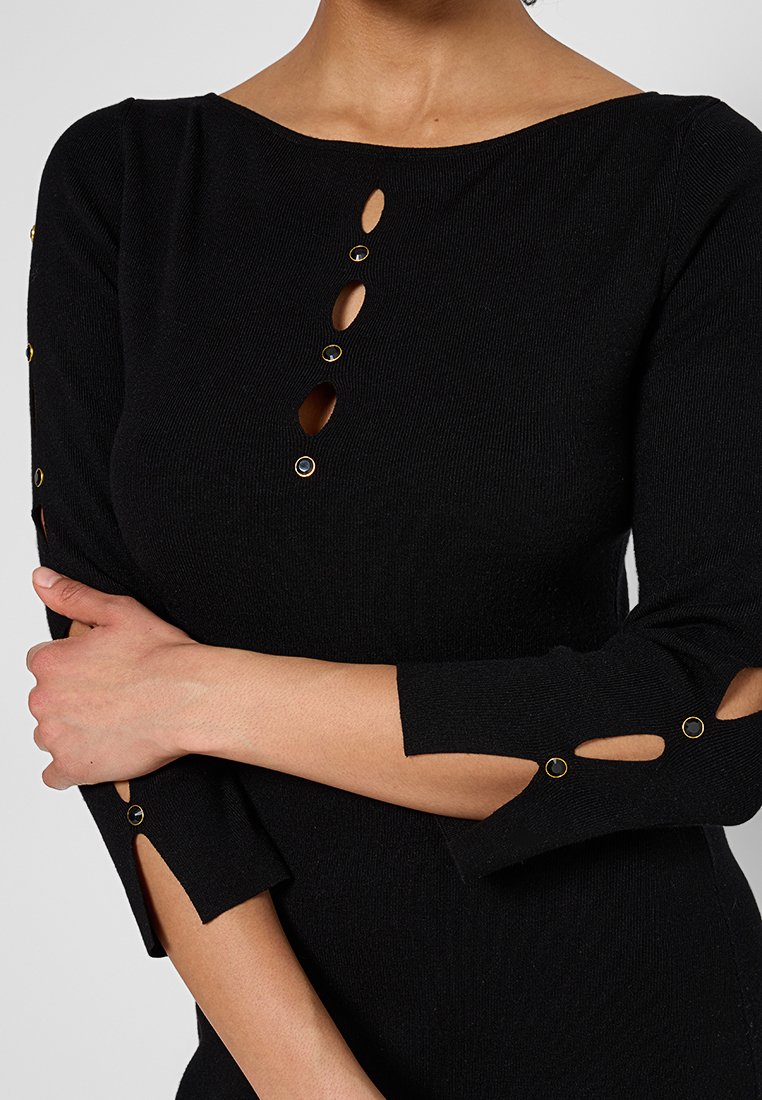 Zwarte geribbelde top met lange mouwen, afgewerkt met gouden accenten, ovale uitsparingen op de mouwen en een rij knopen aan de voorkant.
