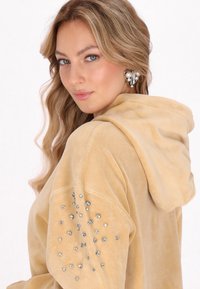 Felpa beige con cappuccio e texture morbida, caratterizzata da accenti di strass disposti su una manica. Design semplice e casual.
