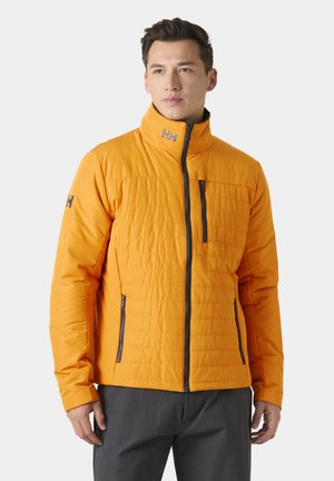 Mężczyzna w jasnopomarańczowej pikowanej kurtce z czarnymi zamkami i logo HH na piersi, kołnierzu oraz lewym rękawie, stojący na jednolitym tle.