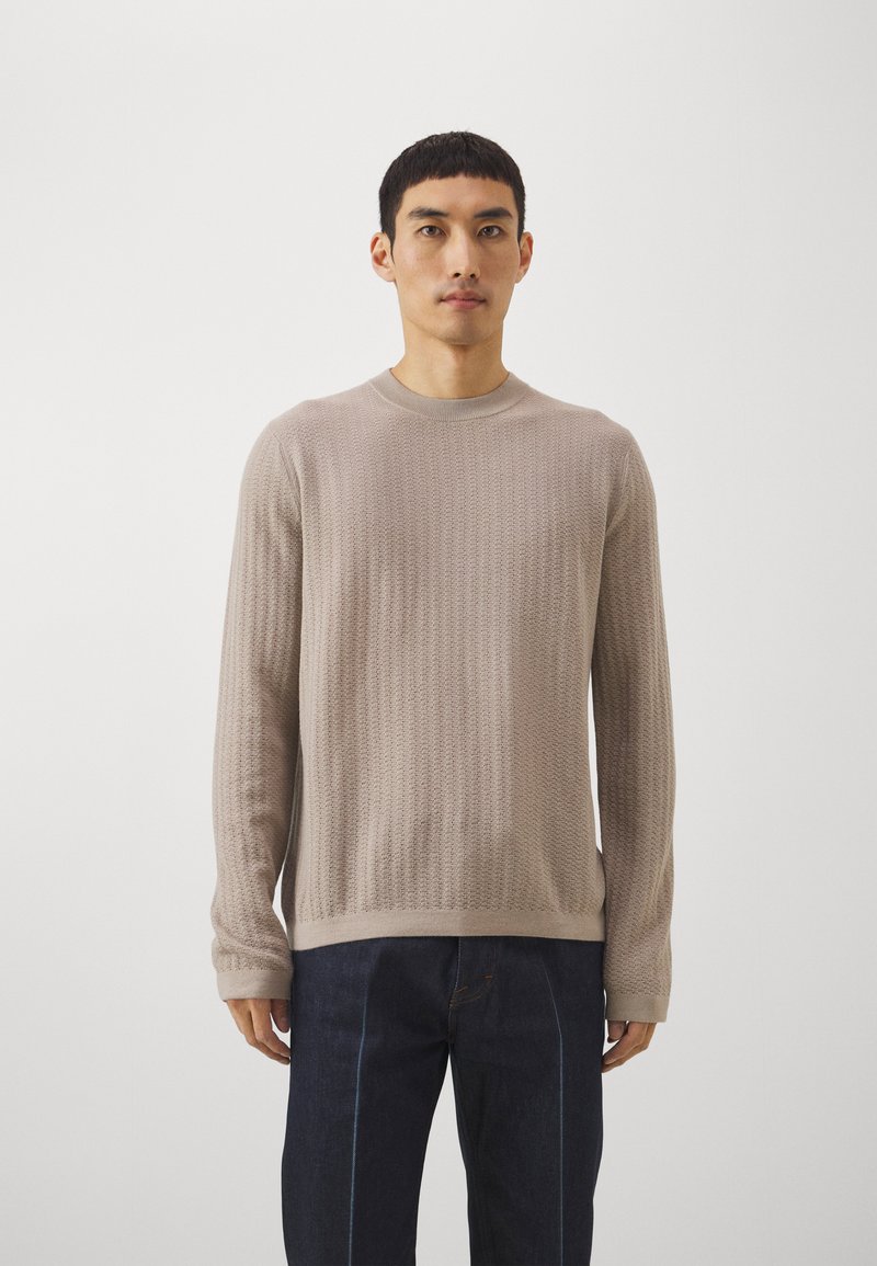 Μπεζ πλεκτό πουλόβερ με στρογγυλή  λαιμόκοψη, υφή και ριγωτές μανσέτες. Φορεμένο με σκούρες τζιν παντελόνες. Απλός, εφαρμοστός σχεδιασμός.