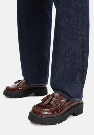 Brązowe mokasyny z tłoczeniem skóry krokodyla, z grubą czarną podeszwą i ozdobami w formie frędzli, zestawione z ciemnoniebieskimi dżinsami o prostych nogawkach.