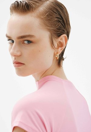 Top ajustado rosa con mangas cortas, textura suave y cuello alto. Arete de aro dorado en la oreja, cabello corto peinado de manera pulida.