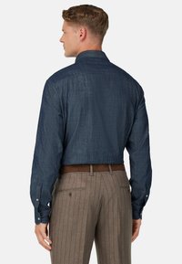 Uomo di spalle che indossa una camicia a maniche lunghe blu navy con texture, infilata in pantaloni marroni a righe con una cintura marrone.