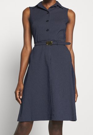 Robe bleu marine sans manches avec petits pois blancs, boutonnée devant, col et ceinture assortie avec boucle dorée à la taille.