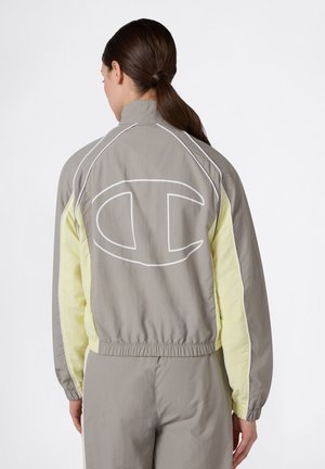 Γυναίκα με σκούρα αλογοουρά που φοράει ένα γκρι και απαλό κίτρινο αδιάβροχο μπουφάν με μεγάλο λευκό περίγραμμα λογότυπου στην πλάτη.