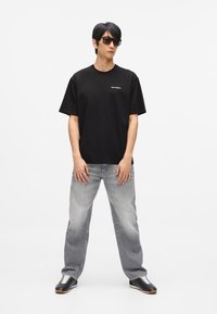 Melna kokvilnas T-kreklu ar īsām piedurknēm un baltu logo uz krūtīm, savienots ar gaiši pelēkiem taisnām kājām džinsiem un melniem apaviem.