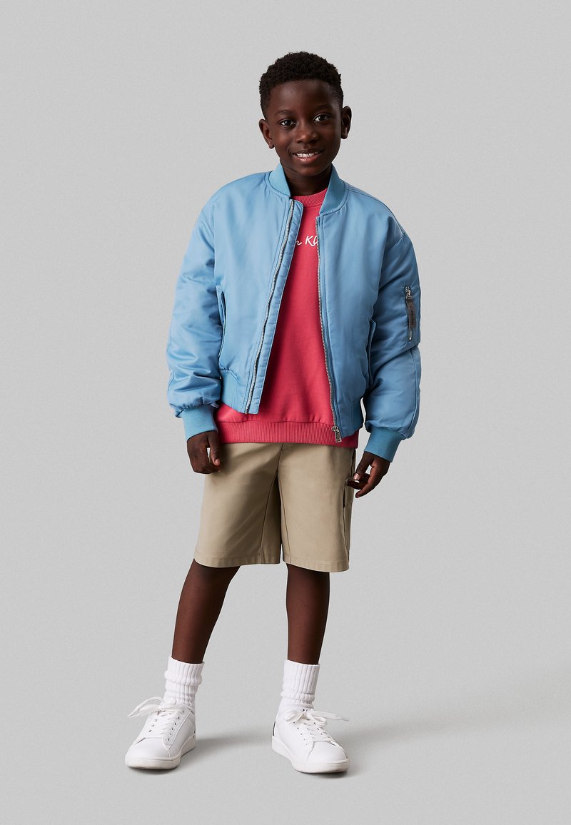 Giacca bomber azzurro chiaro con polsini a coste, felpa rossa sotto, pantaloni corti khaki, sneakers bianche e calzini bianchi alla caviglia.