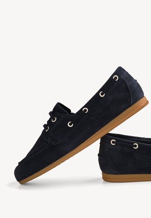 Chaussure bateau en daim bleu foncé avec semelle en caoutchouc beige, fermeture à lacets à l'avant et œillets métalliques, vue de côté sur un fond uni.