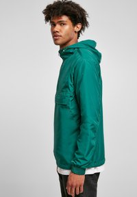 Grøn, letvægts, hættejakke med en frontlomme, elastiske manchetter og en glat, shiny tekstur. Sidevisning med model iført sorte jeans.