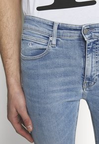 Jeans de mezclilla de color azul claro con un corte regular. La tela tiene una textura desteñida con un ligero desgaste y un pequeño logo en el bolsillo delantero.