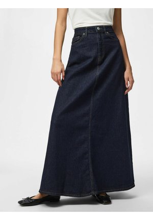 Femme portant une jupe longue en denim bleu foncé et des ballerines noires, debout devant un fond blanc uni.