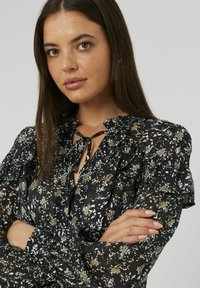 Jeune femme avec de longs cheveux bruns, portant un blouse noire à fleurs avec des volants, les bras croisés, expression neutre, fond gris clair.