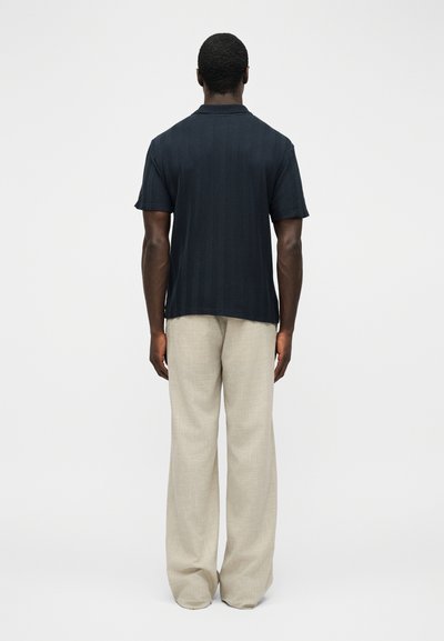 Άνδρας με σκουρόχρωμο δέρμα που φοράει κοντομάνικο μπλε ναυτικό πόλο με υφή και φαρδιά ανοιχτό μπεζ παντελόνια, στέκεται γυρισμένος πλάτη στην κάμερα.