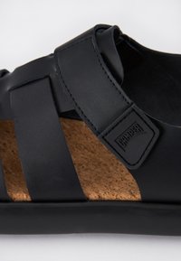 Black sandal featuring wide straps and a cork insole, with a textured finish. Accented by a rubber outsole and a branded label.