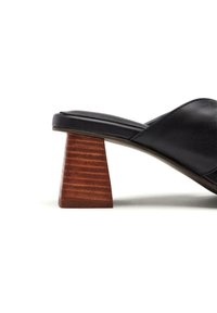 Sandale à glissière en cuir noir avec un talon conique en bois brun texturé. Le design est minimaliste avec une tige lisse et un léger arc.