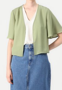 Svetle zelená, krátká otevřená bunda s krátkým rukávem vyrobená z měkkého, splývavého materiálu, přehozená přes bílý top s výstřihem do V a v kombinaci s denimovou sukní.