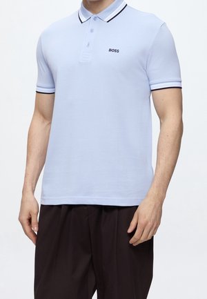 Man draagt een lichtblauw BOSS poloshirt met donkere biezen aan kraag en mouwen, gecombineerd met losse donkere broek, tegen een effen achtergrond.