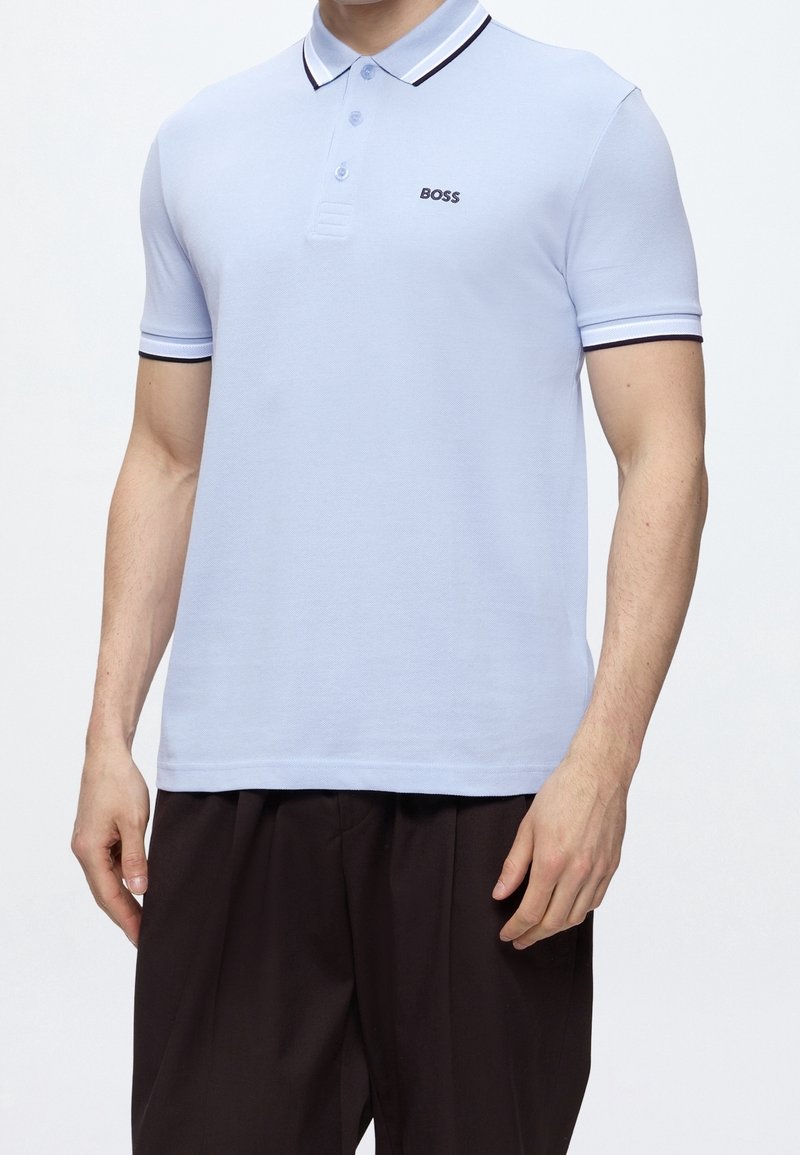 Homme portant un polo BOSS bleu clair avec une bordure foncée au col et aux manches, associé à un pantalon ample foncé, sur un fond uni.