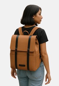 Zaino marrone con finitura opaca, forma rettangolare, dettagli neri, spallacci regolabili e chiusura a linguetta sulla tasca principale.