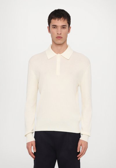 Pull col polo en tricot ivoire avec un col et des manches longues. Caractérisé par une texture douce et une coupe décontractée, associé à un pantalon sombre.