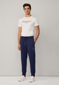 Witte katoenen t-shirt met "HACKETT LONDON" gedrukt in het rood, gecombineerd met marineblauwe joggingbroeken met logodetail en witte sneakers.
