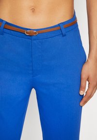 Modré kalhoty vyrobené z hladkého materiálu, obohacené o hnědý kožený pásek s kovovými přezkami. Úzký střih s předními kapsami.