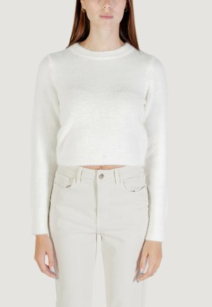 Donna che indossa un maglione corto bianco e peloso e pantaloni chiari beige a vita alta, in piedi di fronte a uno sfondo neutro.