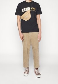 Černé tričko s zlatým logem "CARHARTT", béžové kordové kalhoty, hnědé vysoké tenisky a světlá tan krosbody taška.