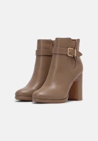 Botas de cuero marrón hasta el tobillo con tacones de bloque, cremallera lateral, correa y detalle de hebilla dorada sobre fondo blanco liso.