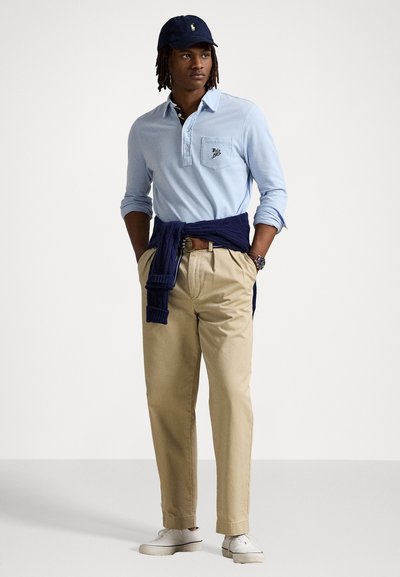 Világoskék, hosszú ujjú póló, mellzsebbel, bézs chinos nadrággal párosítva, derékon kötött navy pulóverrel. A megjelenést fehér sportcipők teszik teljessé.