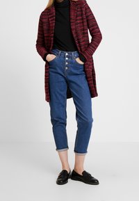 Cardigan a strisce rosse e nere di lunghezza media sopra un dolcevita nero, abbinato a jeans in denim blu a vita alta e mocassini neri.
