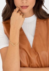 Gold bracelet featuring interlocking circular links and an elongated design, worn on a hand against a brown leather backdrop.