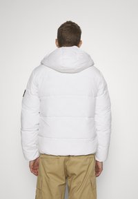 Giacca a vento bianca con cappuccio, caratterizzata da segmenti trapuntati e una texture liscia. I dettagli includono un'etichetta nera sulla manica sinistra.