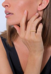 Goldene, gedrehte Ring, der am Zeigefinger getragen wird, mit einem strukturierten Design. Die Hand des Models berührt sanft ihr Gesicht und zeigt die Details des Rings.