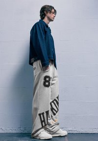 Fiatal személy áll oldalra fordulva, kék farmerdzsekiben, szürke bő nadrágban fekete felirattal és fehér sportcipőben, egy világos fal előtt.